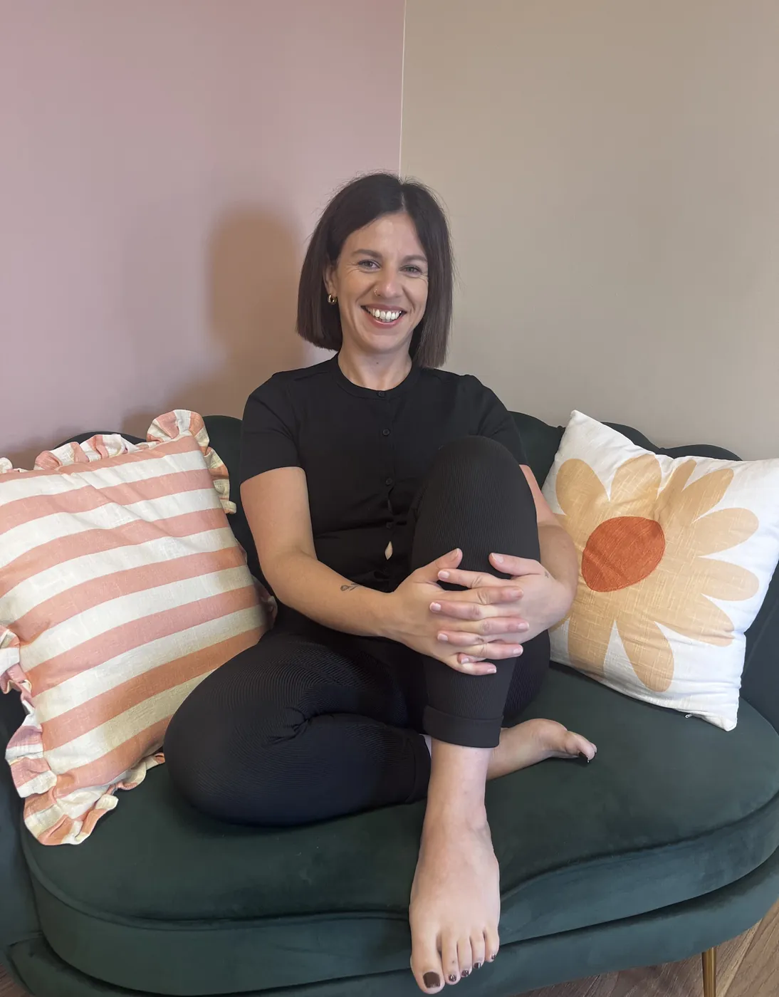 Lucie, souriante, assise pieds nus sur le canapé de velours vert.