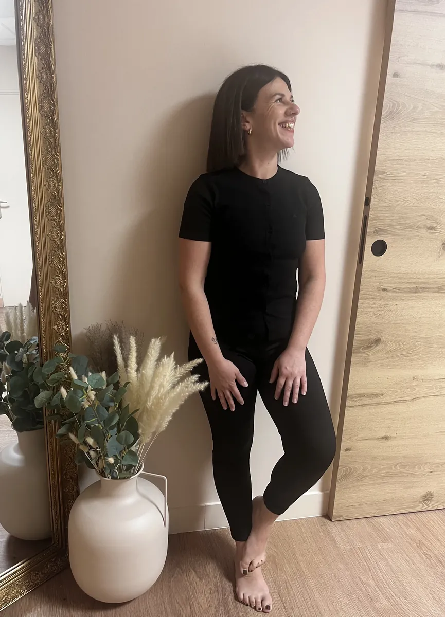 Lucie, debout contre le mur du salon à côté d'un grand miroir doré, sourit en regardant au loin.